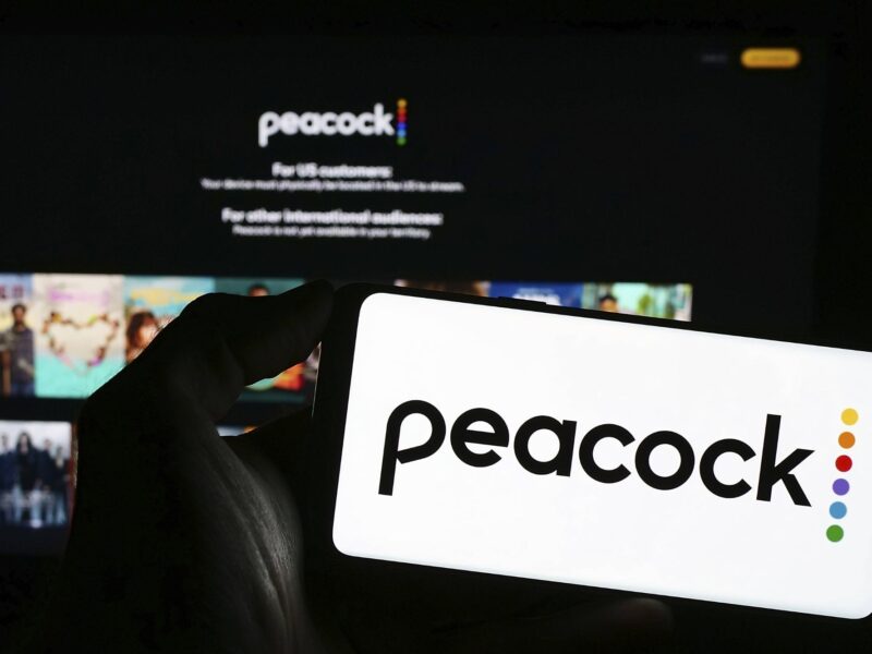 The Peacock logo being displayed on a mobile phone screen in front of a TV with the Peacock app playing.
